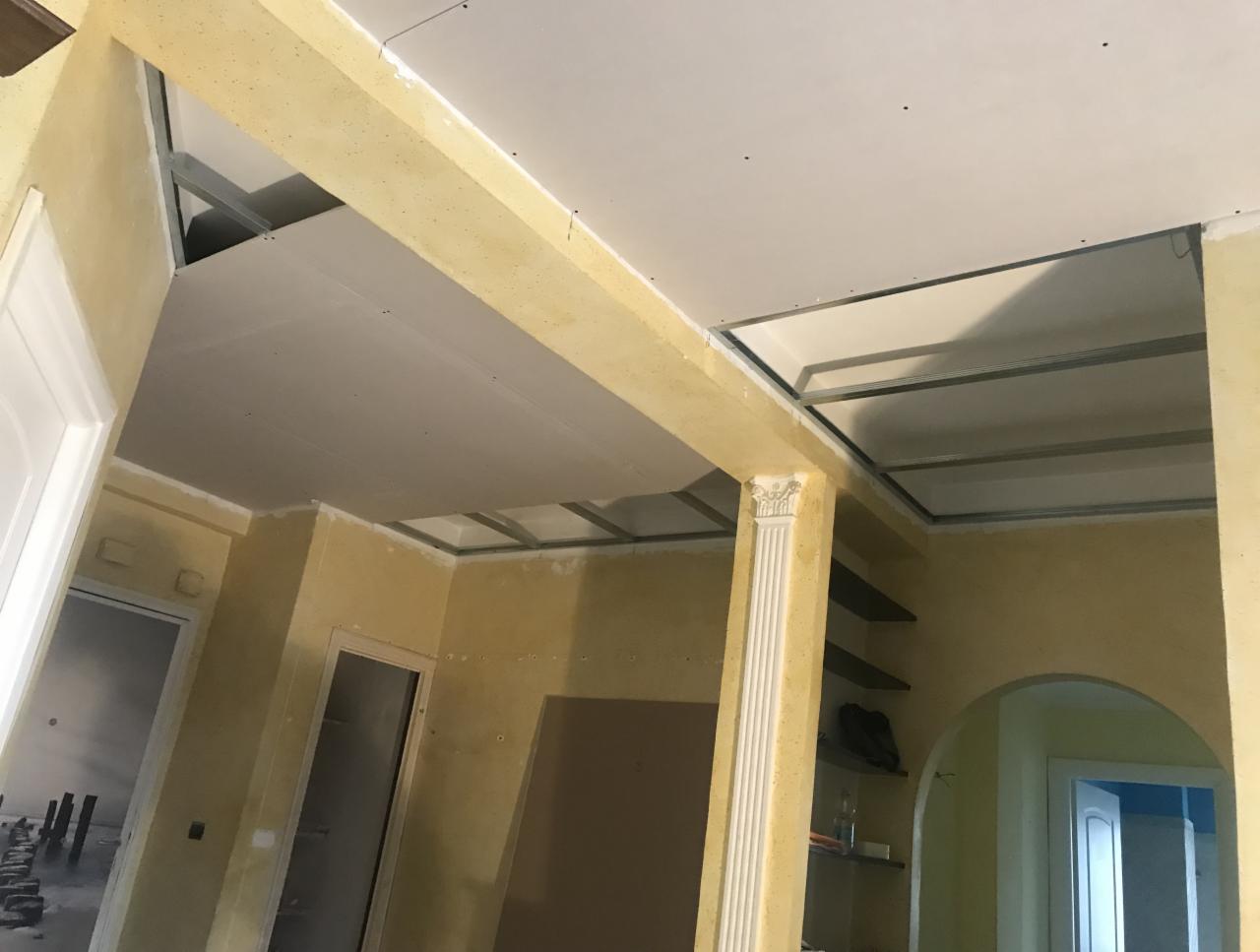 Wnętrze w trakcie montażu podwieszanego sufitu z płyt gipsowo-kartonowych, widoczne metalowe profile mocujące, żółte ściany z widocznymi niedoskonałościami faktury, kolumna dekoracyjna i fragmenty...