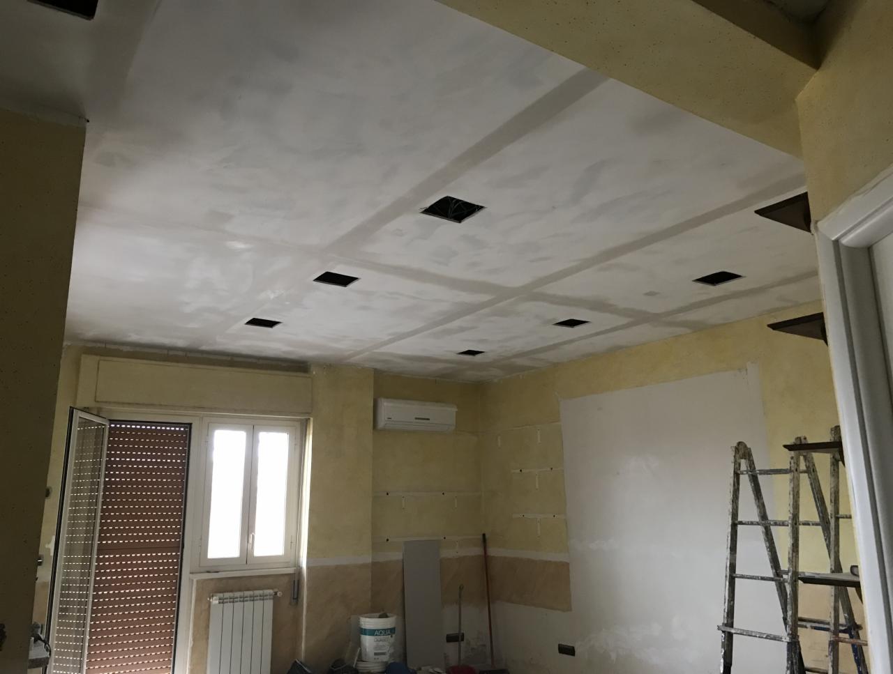Pokój w trakcie prac wykończeniowych: sufit z nałożoną gładzią i przygotowanymi otworami na oświetlenie, ściany częściowo pomalowane na żółto, widoczna drabina i wiadro z farbą.