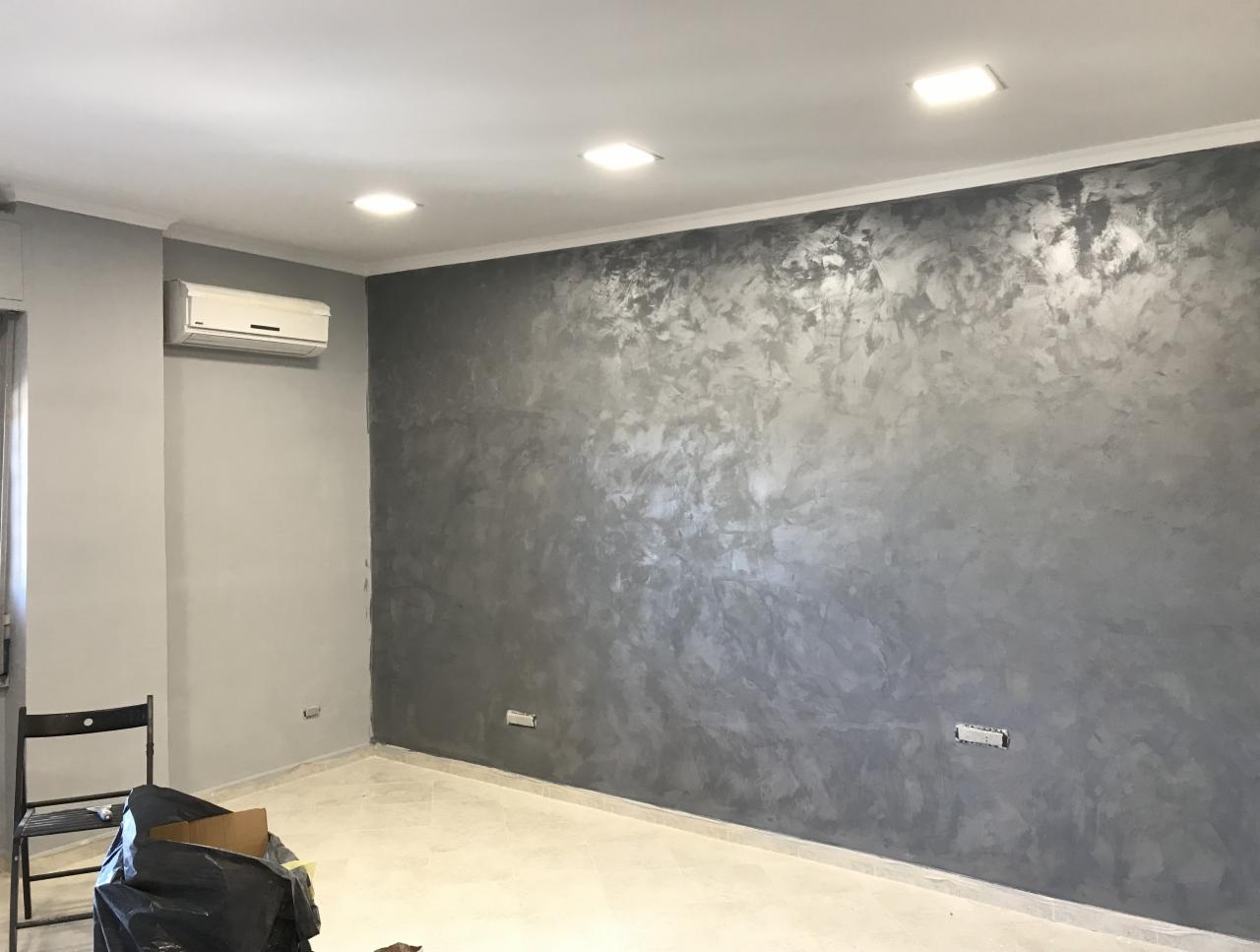 Ściana w pokoju po nałożeniu dekoracyjnej, szarej gładzi w stylu weneckim, widoczny klimatyzator na sąsiedniej ścianie oraz przygotowane do uprzątnięcia odpady budowlane.