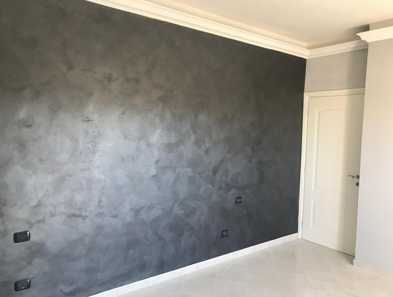 Ściana w pokoju pokryta dekoracyjną, ciemnoszarą farbą imitującą efekt betonu, z widocznymi włącznikami światła i białą listwą przypodłogową, obok białe drzwi.