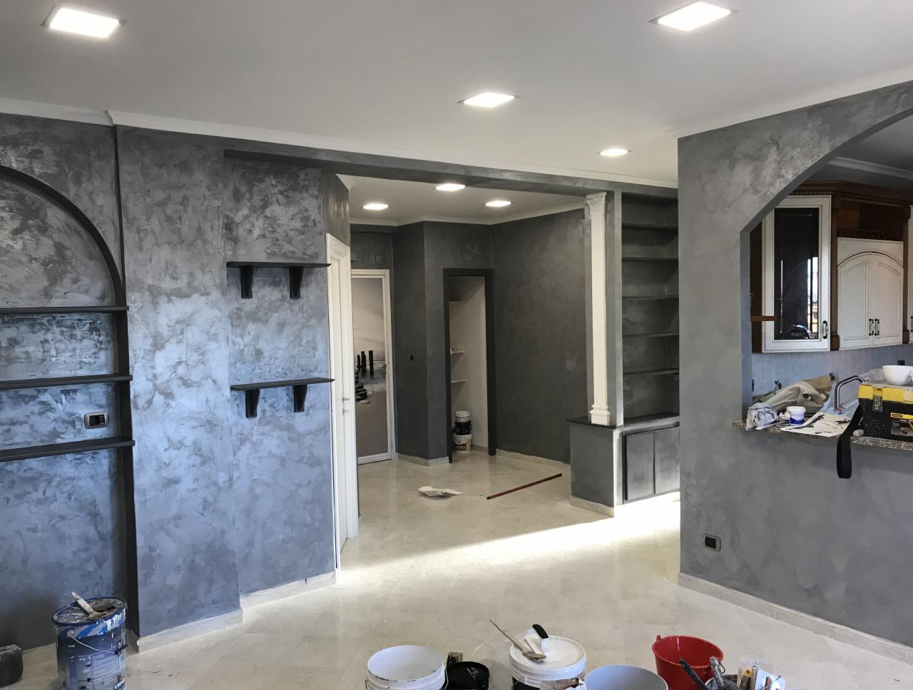 Wnętrze po nałożeniu dekoracyjnej, szarej gładzi na ścianach, widoczne pędzle, wałki i wiadra z farbą na podłodze, wnętrze w trakcie prac wykończeniowych.