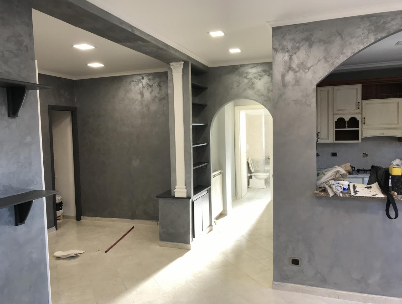 Wnętrze po malowaniu dekoracyjną farbą w odcieniach szarości, z widocznym fragmentem kuchni i łazienki w tle, podkreślone sztucznym oświetleniem punktowym.