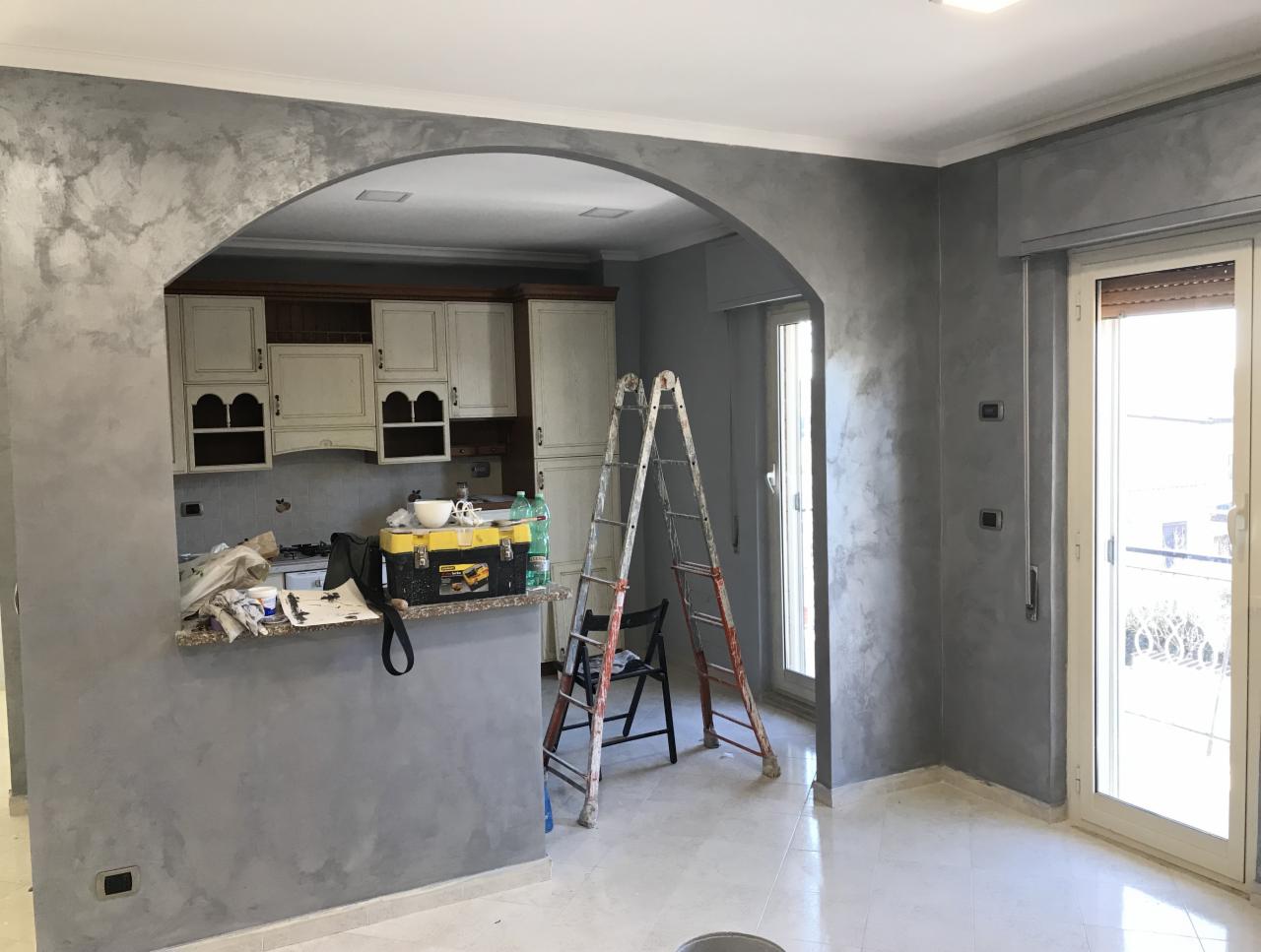 Wnętrze mieszkania w trakcie prac wykończeniowych: ściany pokryte szarą, dekoracyjną farbą z efektem cieniowania, widoczna drabina i narzędzia malarskie, kuchnia z białymi szafkami w tle, marmurowe...