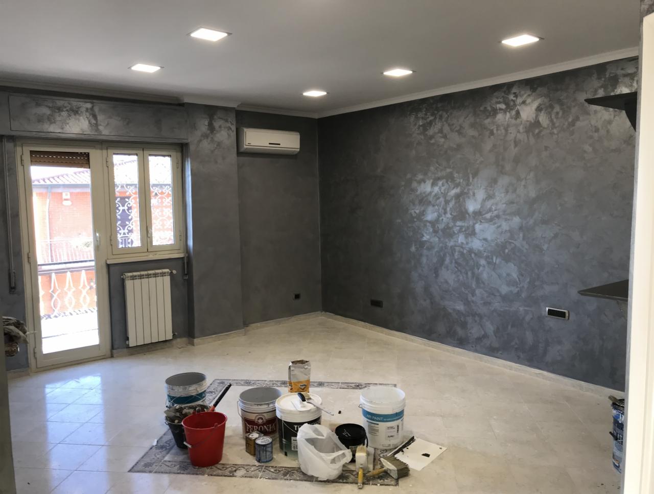 Pokój po nałożeniu dekoracyjnej, szarej farby ze srebrnymi refleksami na ścianach; widoczne puszki z farbą na podłodze, grzejnik pod oknem balkonowym.