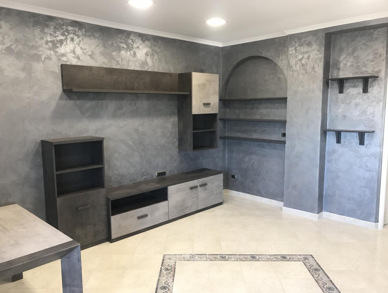 Szara ściana w salonie wykończona dekoracyjną techniką malarską imitującą beton, z zabudową meblową w odcieniach szarości i beżu, podłoga z jasnych płytek z dekoracyjnym obramowaniem.