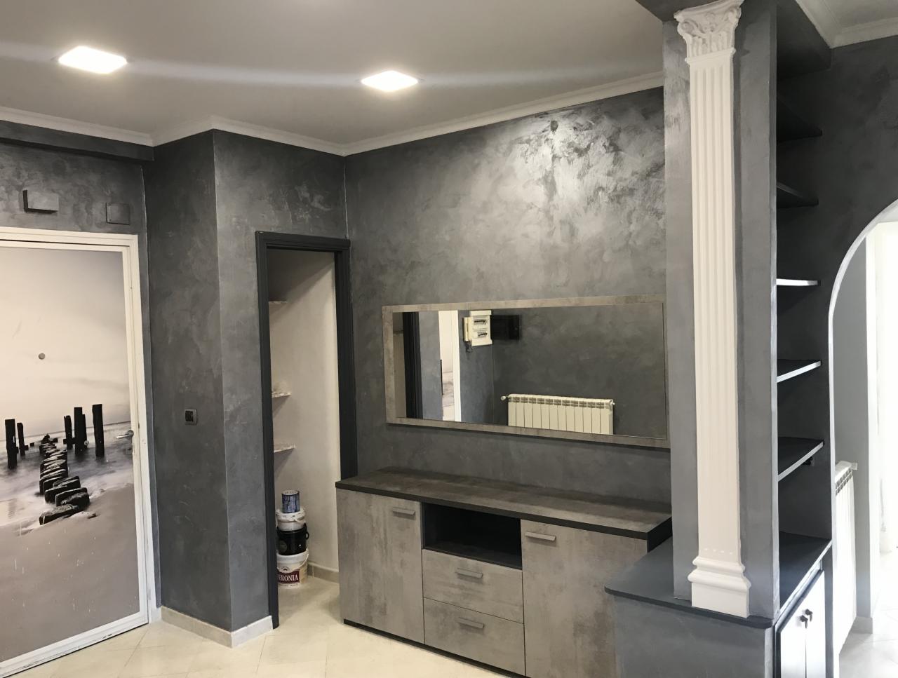 Pokój z szarymi ścianami wykończonymi efektem dekoracyjnym, widoczne lustro nad szafką, drzwi z fototapetą i dekoracyjny filar.