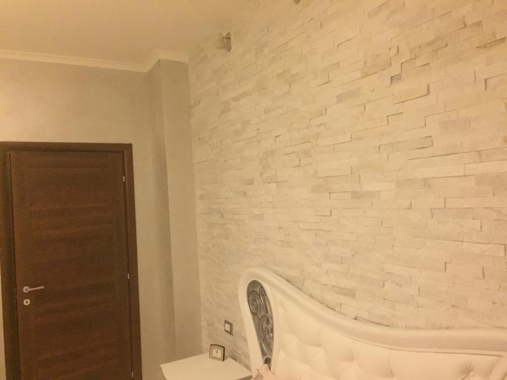 Ściana sypialni wykończona dekoracyjną okładziną z białego kamienia, widoczna część łóżka z ozdobnym zagłówkiem i fragment drzwi w odcieniach brązu.