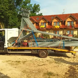 Biały bus z otwartymi drzwiami kierowcy przewożący konstrukcje metalowe i agregat prądotwórczy, zabezpieczone pasami transportowymi, na tle budynku z czerwonym dachem.
