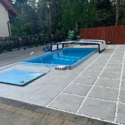 Nowoczesna aranżacja przestrzeni wokół basenu z szarych płyt w kwadraty, wypełnionych białym grysem. W tle zieleń drzew i częściowo widoczny budynek.