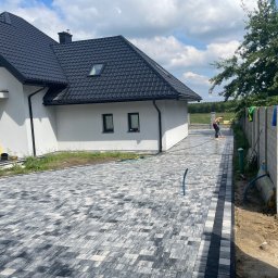 CHOJAK-BRUK - Nowoczesny podjazd z szarej kostki brukowej ułożonej we wzór, obok biały dom z czarnym dachem i pracownik zamiatający powierzchnię. Widok na ogrodzenie i zieleń.