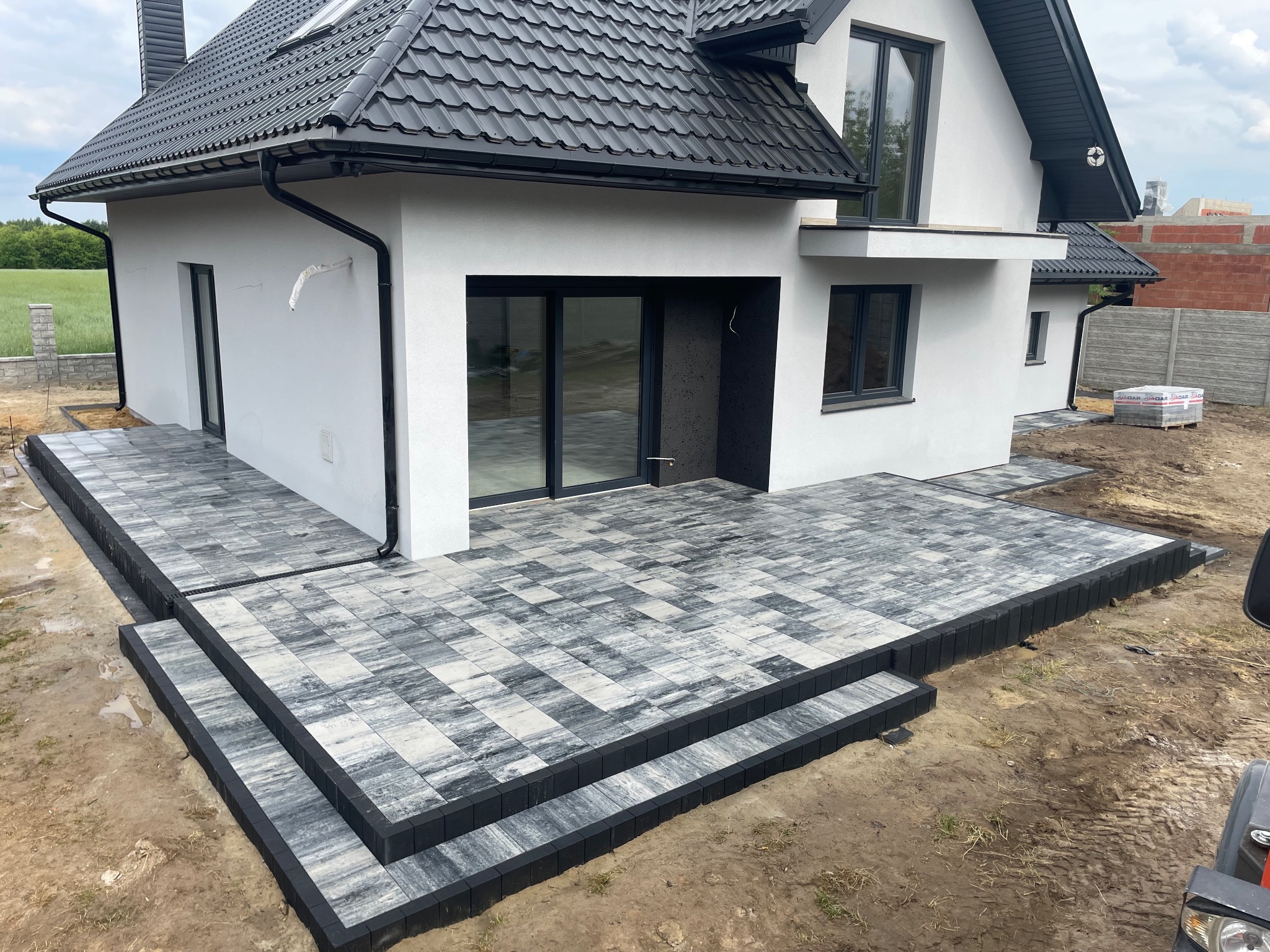 Nowoczesny taras z kostki brukowej w Ciepielowie. Schody z szarej kostki prowadzą do jasnego domu z czarnym dachem. Estetyczne wykończenie.