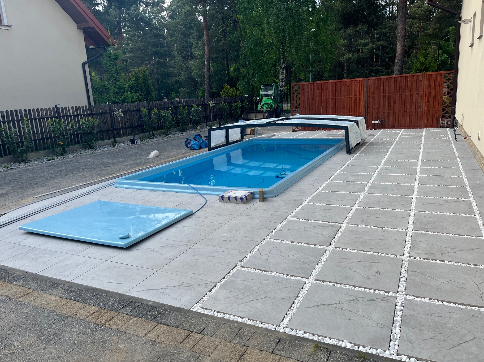 Nowoczesna aranżacja przestrzeni wokół basenu z szarych płyt w kwadraty, wypełnionych białym grysem. W tle zieleń drzew i częściowo widoczny budynek.