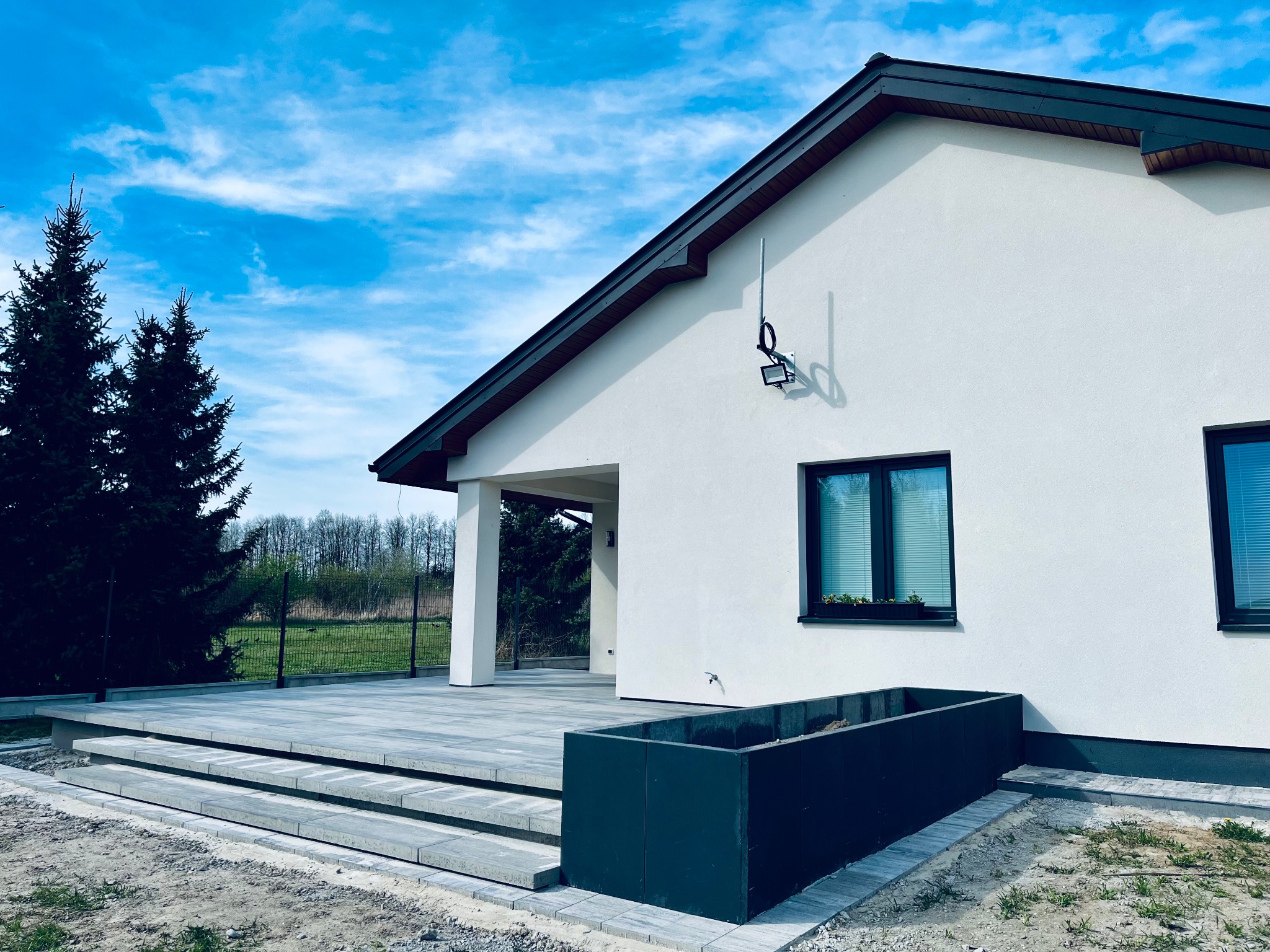 Nowoczesny dom z tarasem wyłożonym szarą kostką brukową. Schody z kostki prowadzą na taras. Minimalistyczna donica wzdłuż ściany.