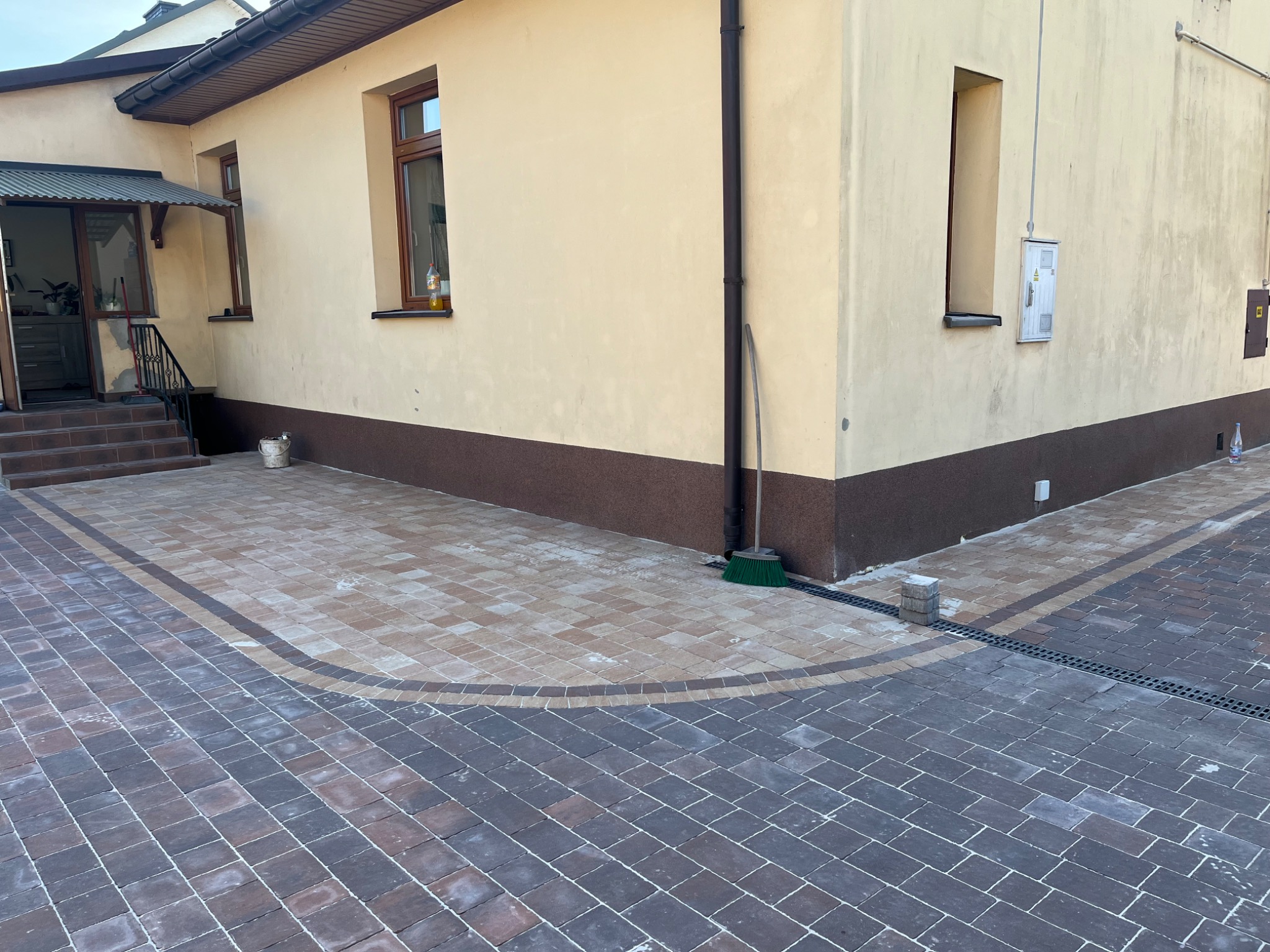 Ułożona kostka brukowa w dwóch odcieniach z dekoracyjnym obramowaniem. Widoczne odwodnienie liniowe i fragment budynku z beżową elewacją i brązowym cokołem.