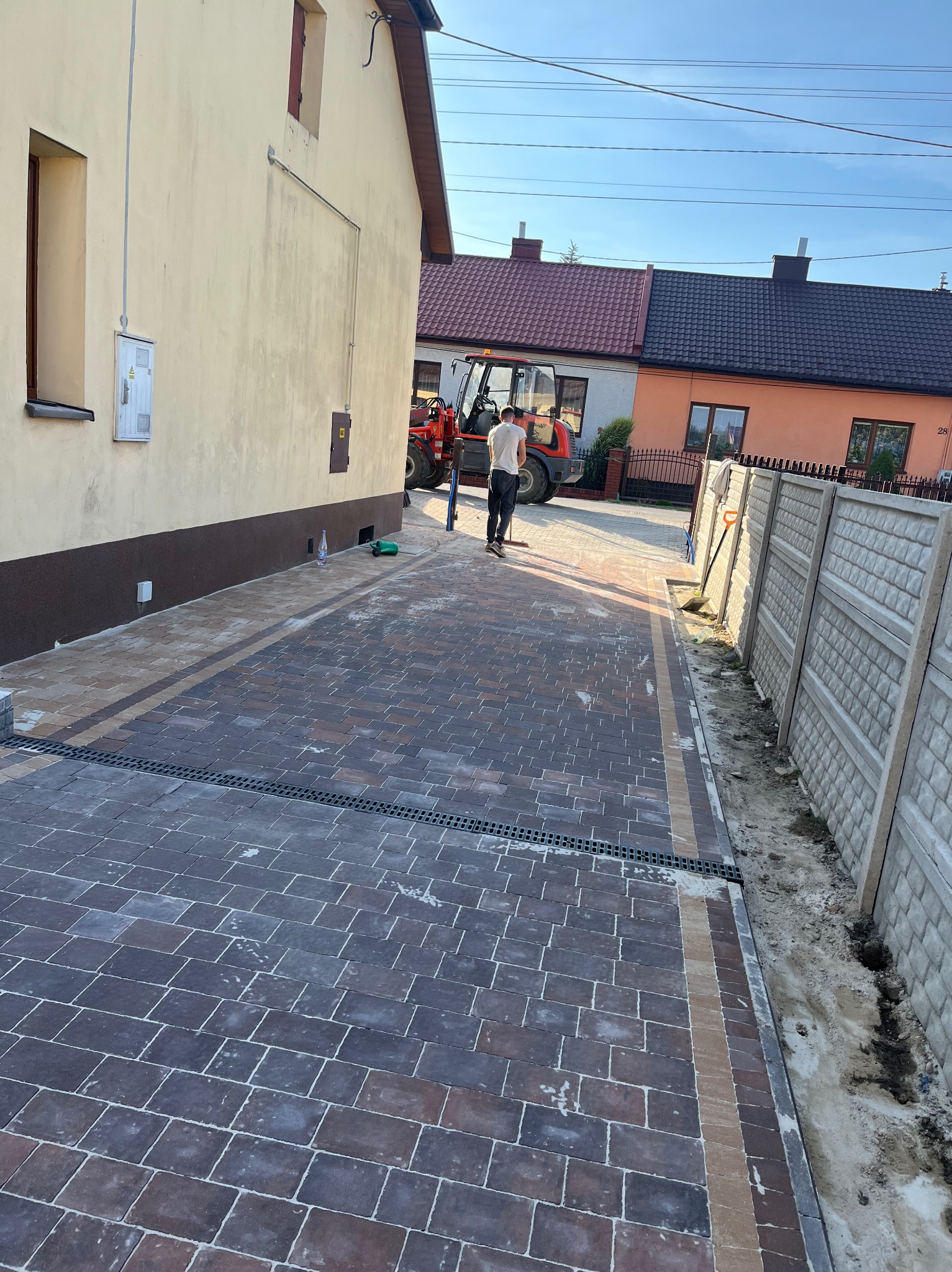 Ukończony podjazd z kostki brukowej w odcieniach brązu i szarości z odpływem liniowym, obok budynku i częściowo zamontowanego ogrodzenia. W tle maszyna budowlana.