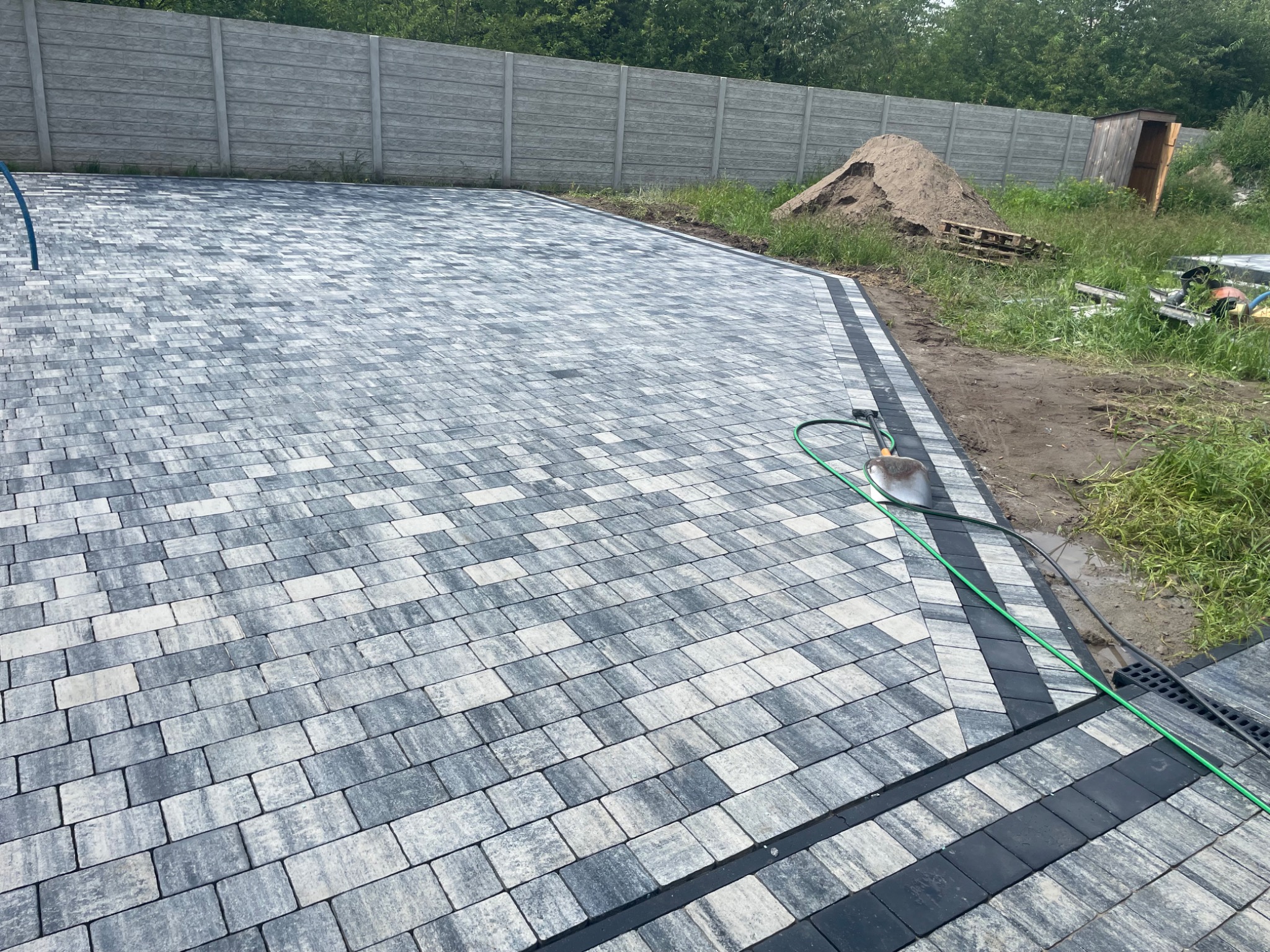 Ułożona kostka brukowa w odcieniach szarości i bieli, z czarnym obramowaniem. W tle betonowe ogrodzenie, ziemia i trawa. Widoczny wąż ogrodowy.