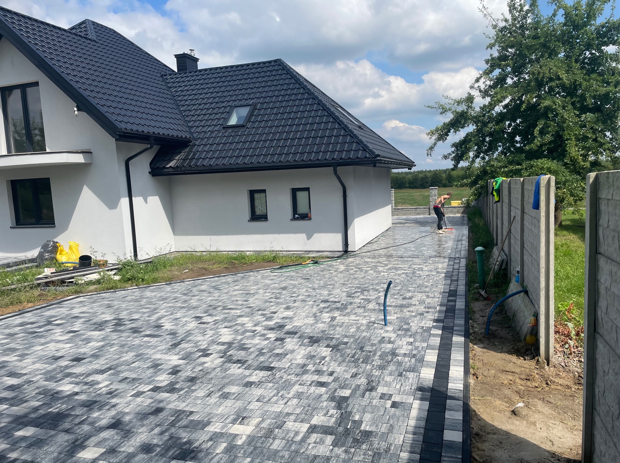 Nowoczesny podjazd z szarej kostki brukowej ułożonej we wzór, obok biały dom z czarnym dachem i pracownik zamiatający powierzchnię. Widok na ogrodzenie i zieleń.