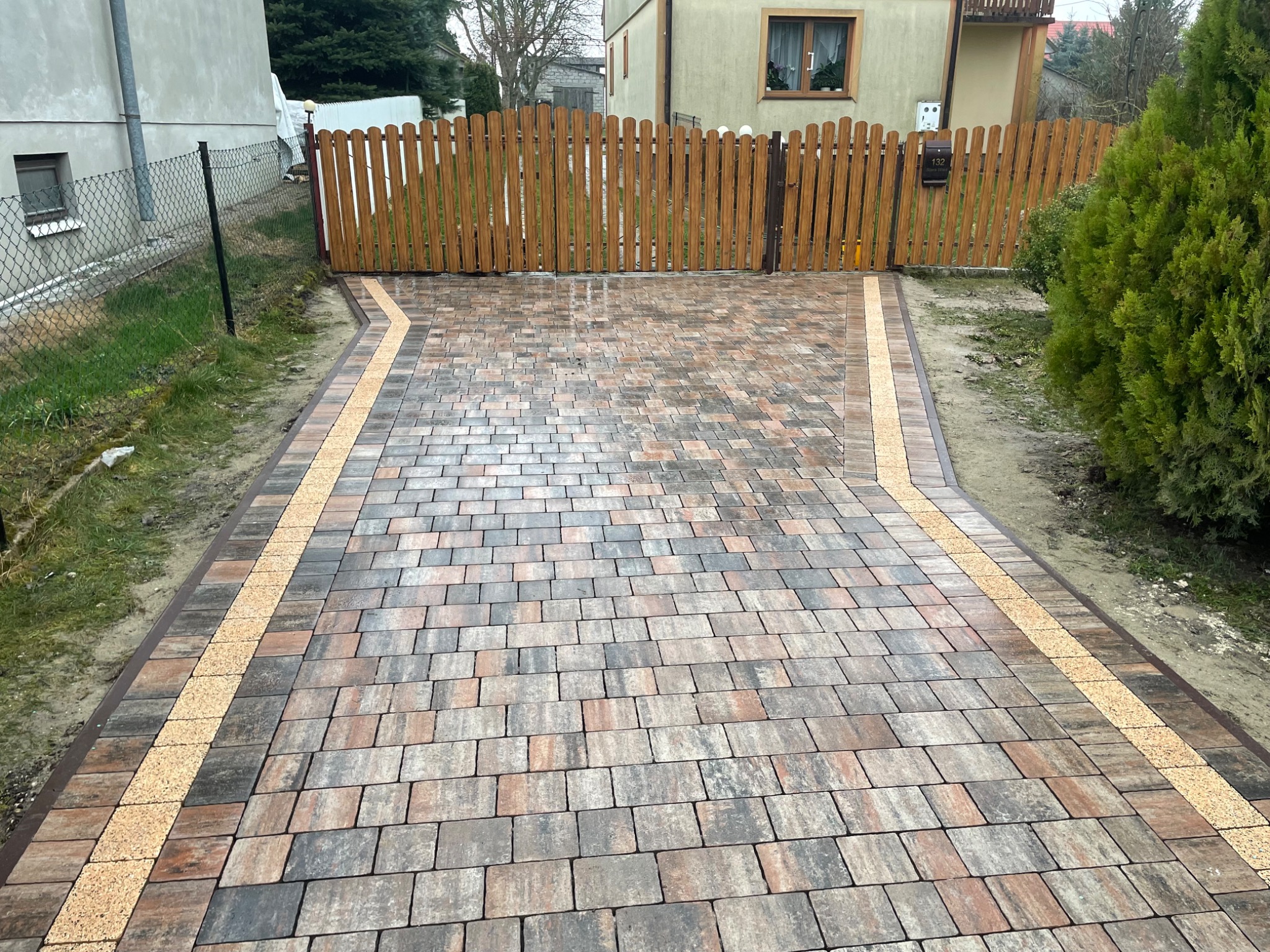 Kolorowa kostka brukowa na podjeździe, obrzeża z jasnego kruszywa, drewniany płot z numerem domu w tle, zieleń po bokach. Widok z perspektywy.