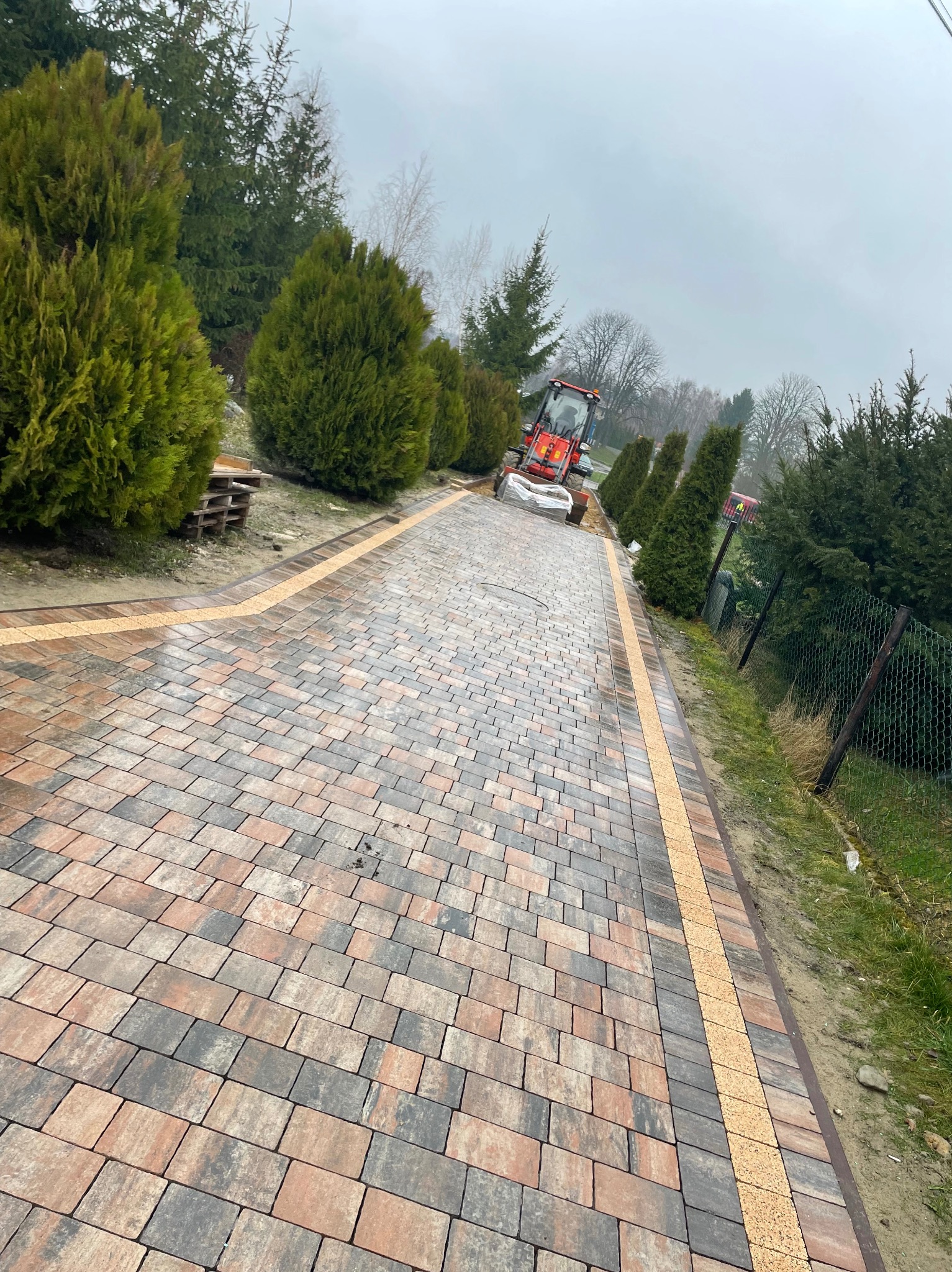 Ukończony podjazd z kostki brukowej w odcieniach szarości i brązu, obrzeża z jasnej kostki. W tle maszyna do układania kostki i zielone tuje.