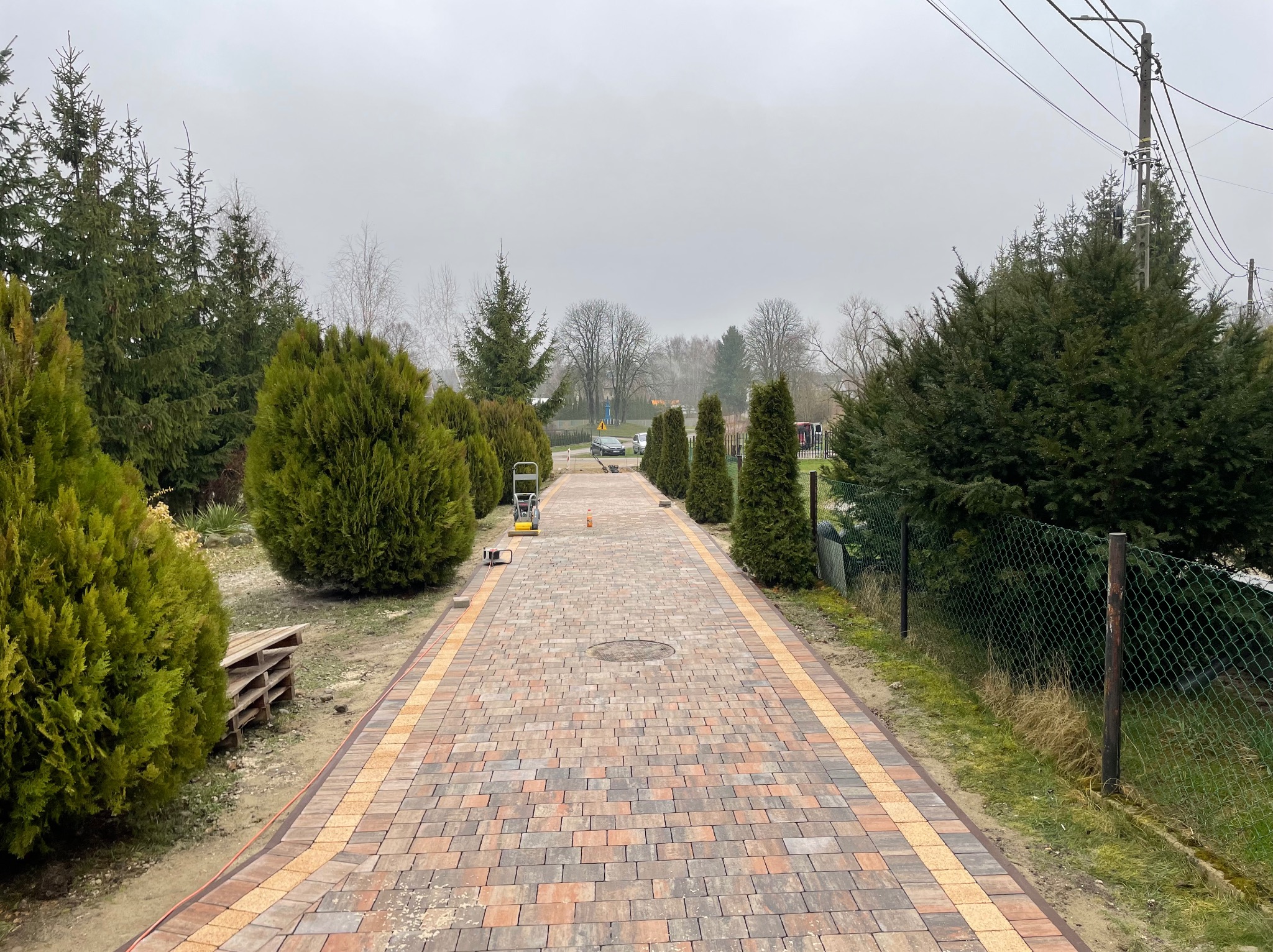 Długa, nowa ścieżka z kostki brukowej w odcieniach brązu i szarości, otoczona zielonymi tujami i drzewami. W tle widoczny sprzęt budowlany i zamglone niebo.