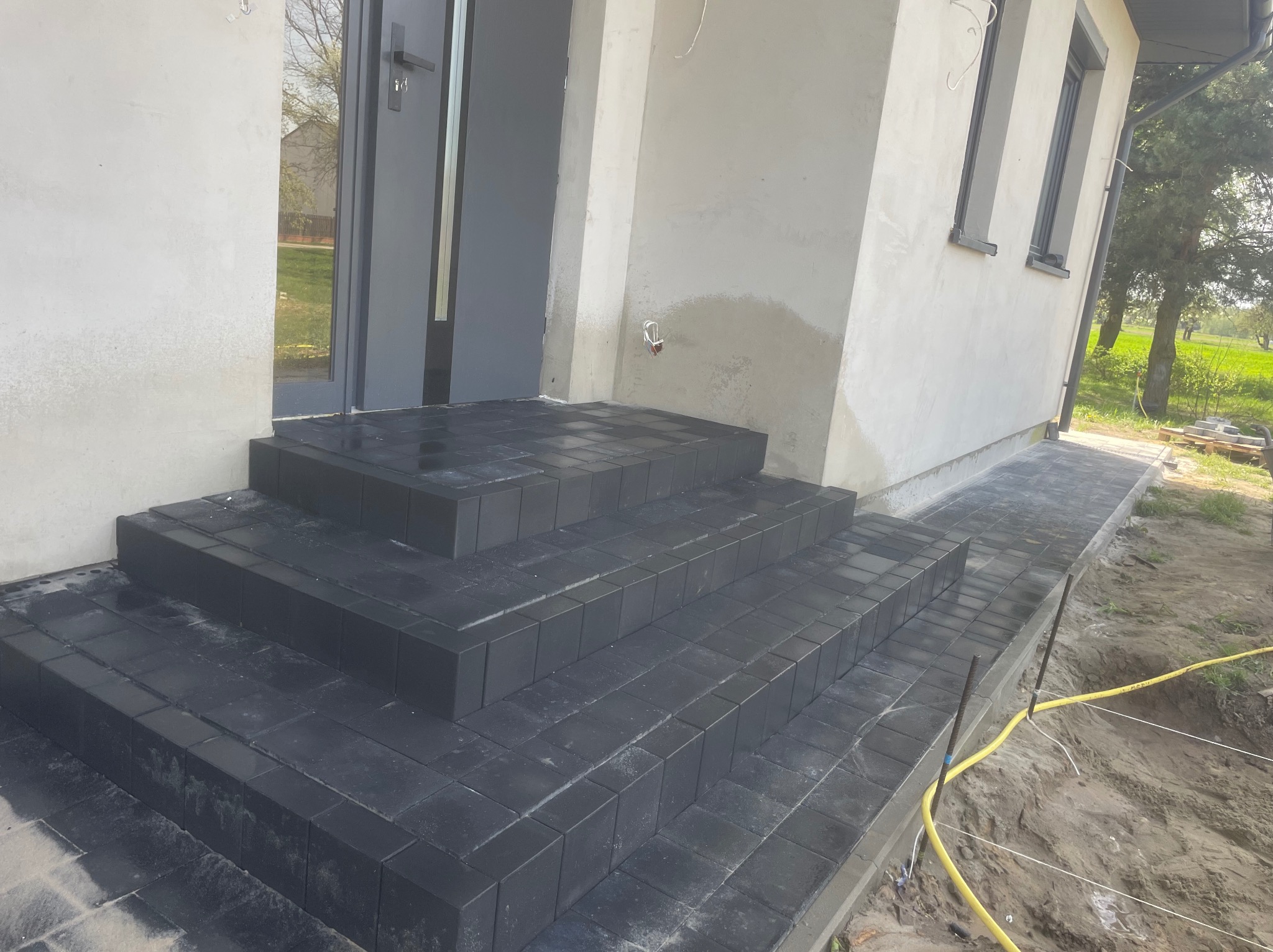 Czarne, kwadratowe płytki tworzą schody wejściowe do domu. Widoczny fragment chodnika i elewacja budynku w jasnym kolorze. Prace brukarskie w toku.
