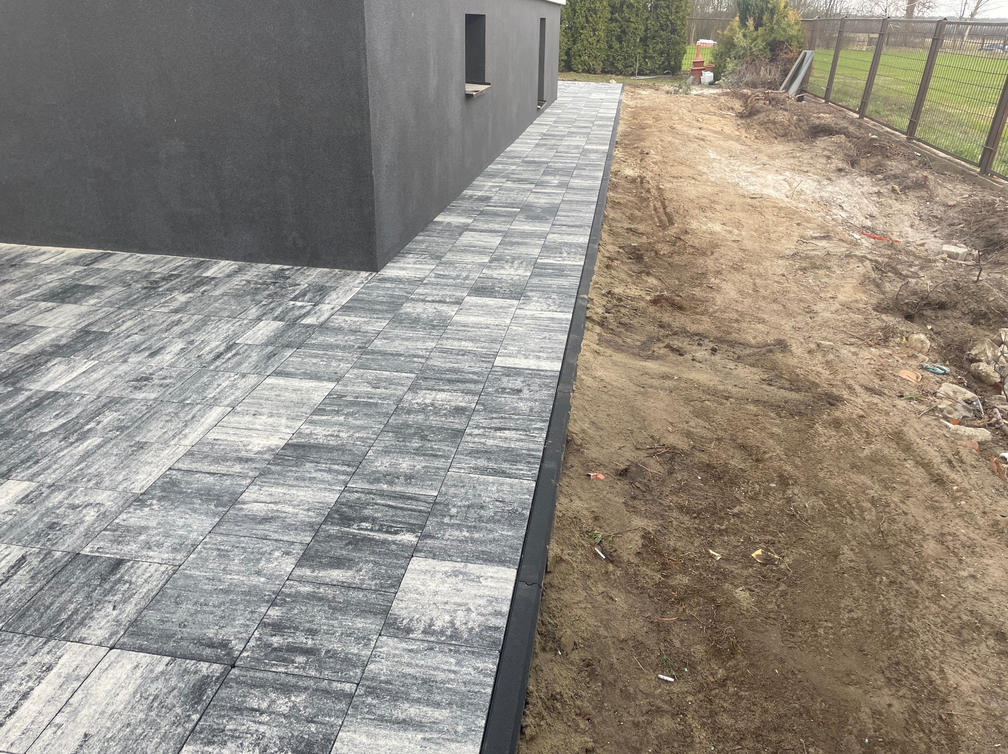 Ułożona ścieżka z szarej kostki brukowej prowadząca wzdłuż ciemnoszarego budynku, obok ziemia. Widok z góry, lekki kąt.