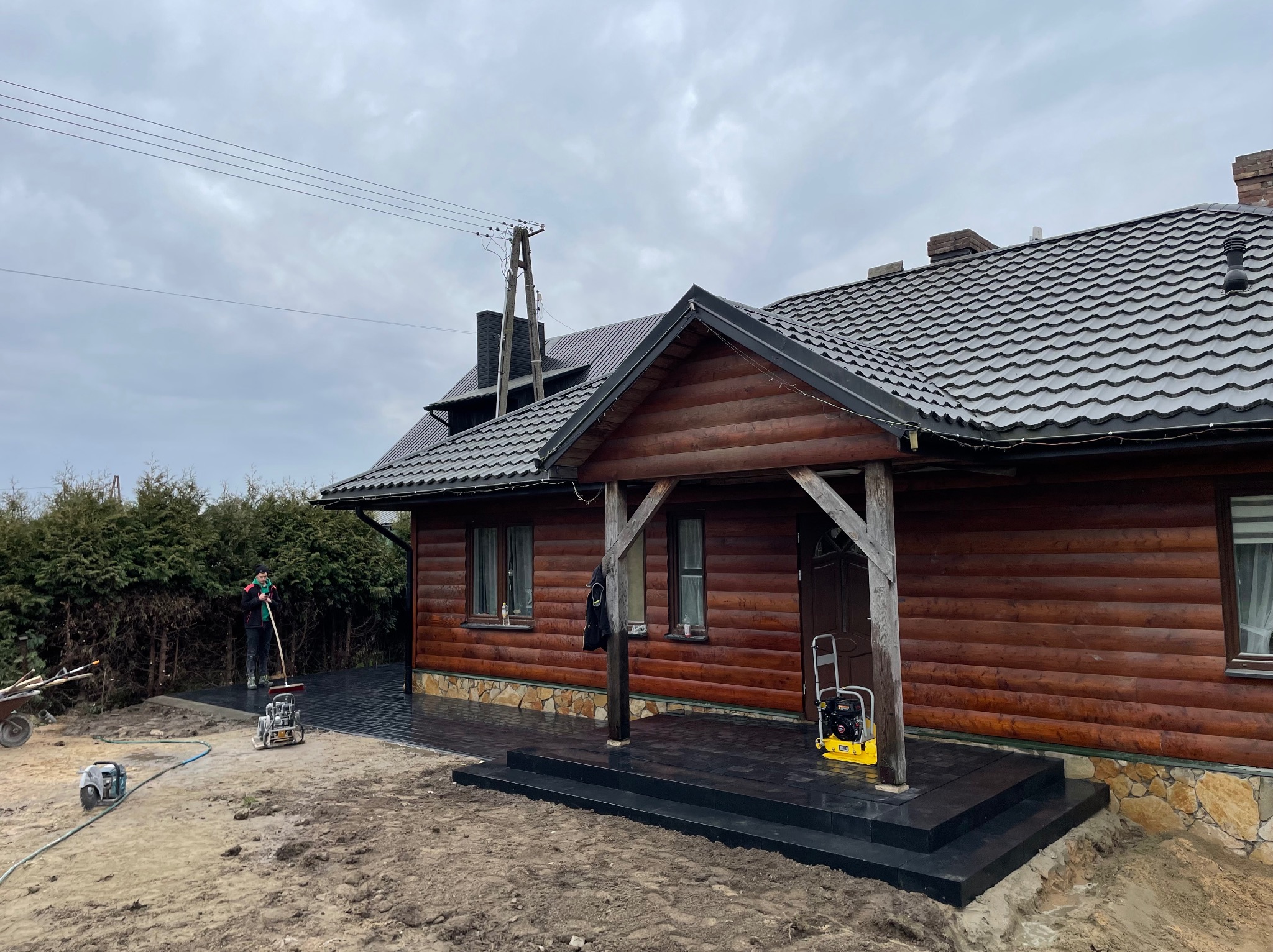 Ukończona nawierzchnia z czarnej kostki brukowej przed domem z bali, z widocznymi schodami wejściowymi. Pracownik w tle zamiata powierzchnię.