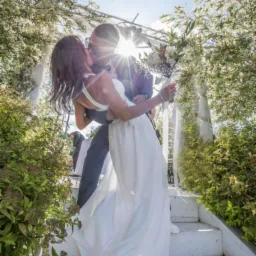 Pocałunek nowożeńców na białych schodach pod kwitnącą pergolą, z promieniami słońca przesłaniającymi ich twarze.