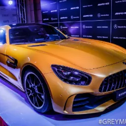 Złoty samoch&oacute;d Mercedes-Benz AMG GT na podwyższeniu z logo marek Ciroc i Grupa Wr&oacute;bel w tle, oświetlony fioletowym światłem.