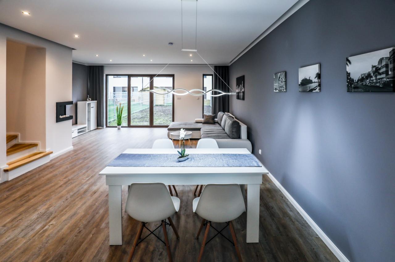 Jasne, przestronne wnętrze domu z drewnianą podłogą, białym stołem jadalnym z niebieskim bieżnikiem, szarą sofą narożną i nowoczesnym oświetleniem.