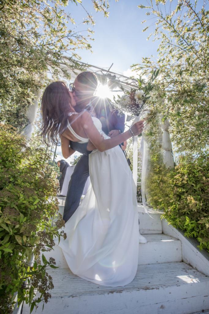 Pocałunek nowożeńców na białych schodach pod kwitnącą pergolą, z promieniami słońca przesłaniającymi ich twarze.