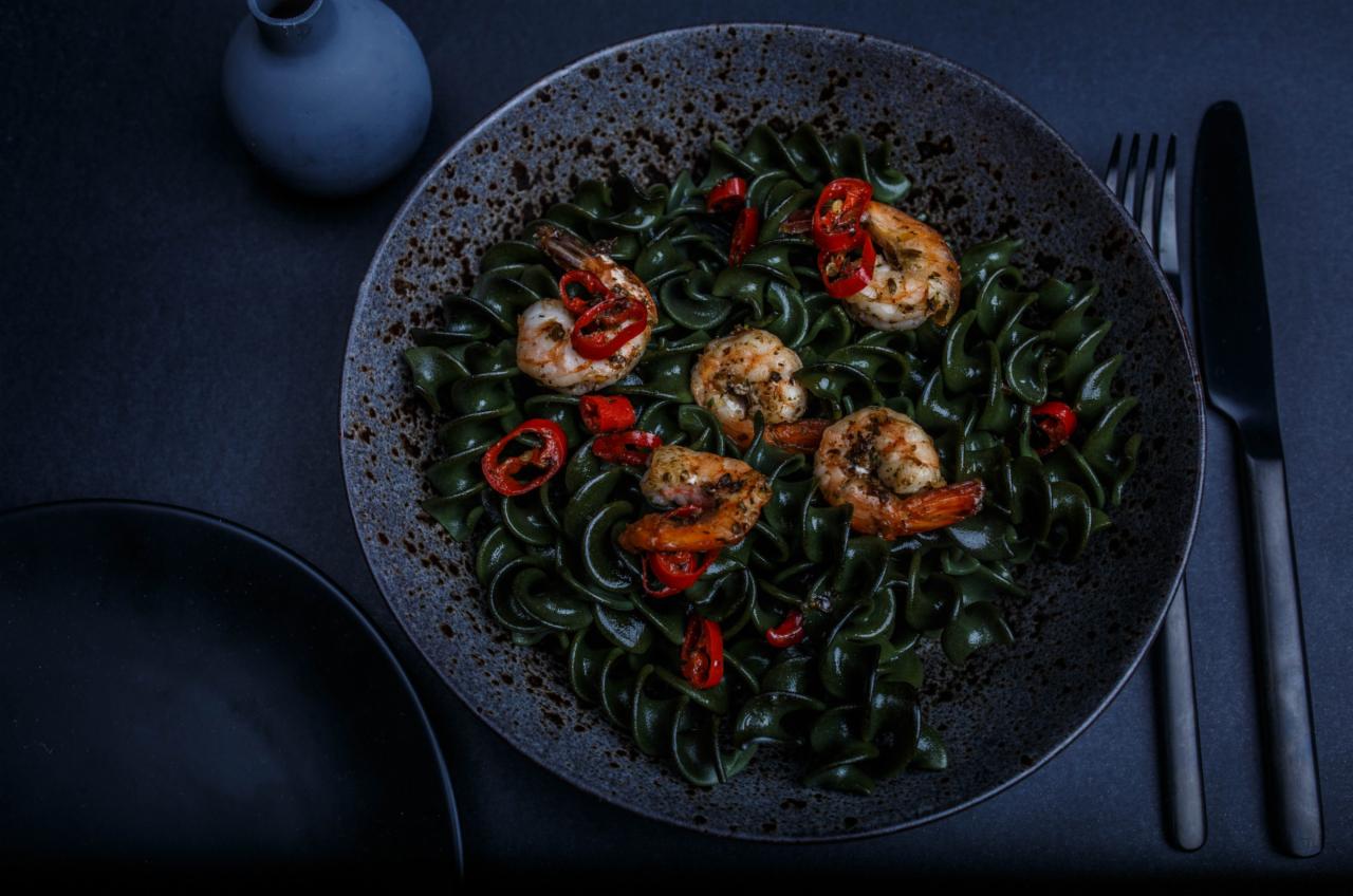 Czarny talerz z zielonym makaronem, krewetkami i papryczką chili na ciemnym tle, obok czarne sztućce i ceramiczny wazon.