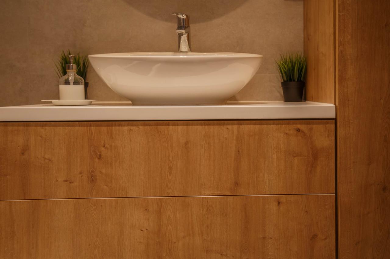 Nowoczesna łazienka z białą umywalką nablatową, drewnianymi szafkami, minimalistyczną baterią i dodatkami w postaci dozownika mydła i małych roślin.