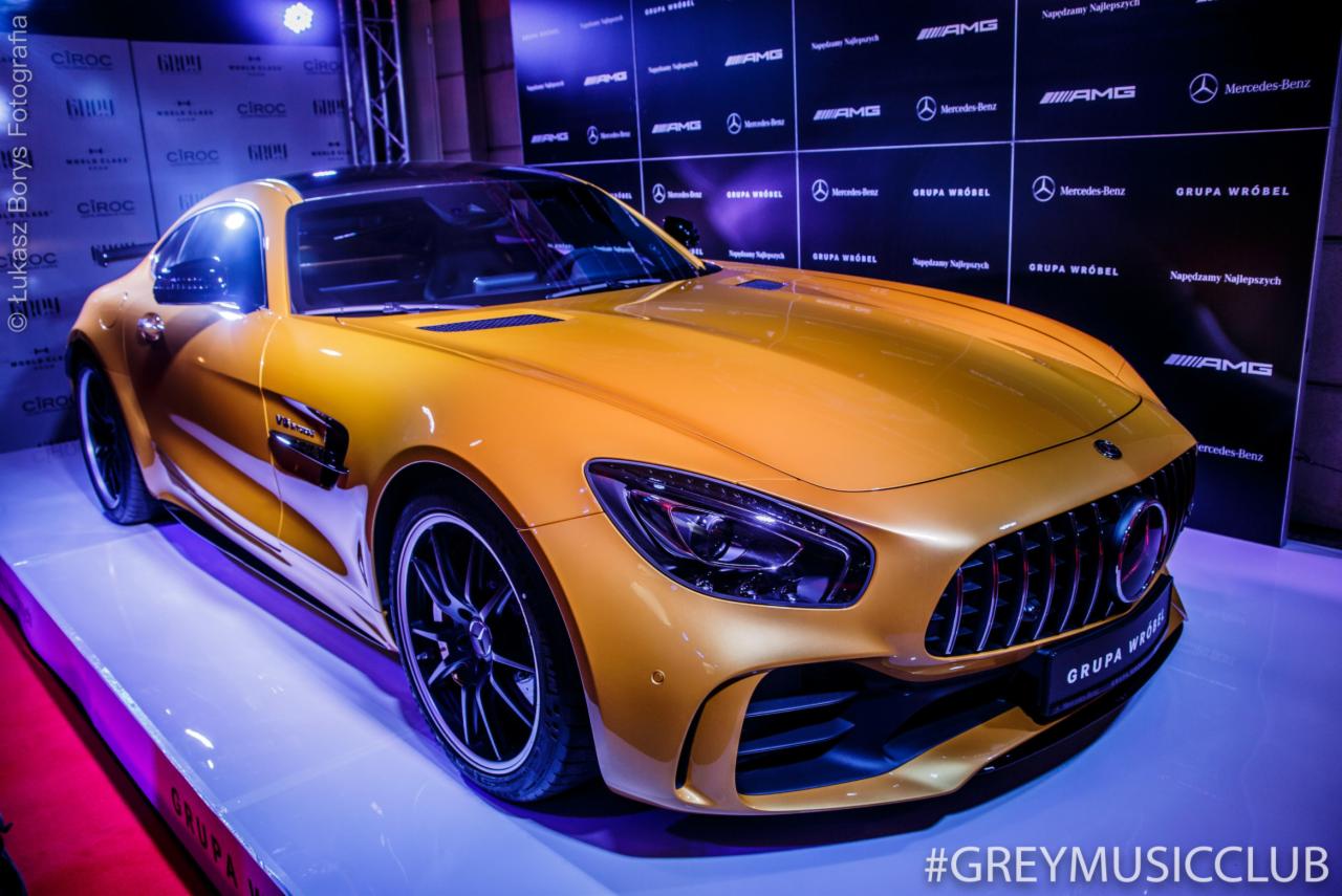 Złoty samochód Mercedes-Benz AMG GT na podwyższeniu z logo marek Ciroc i Grupa Wróbel w tle, oświetlony fioletowym światłem.