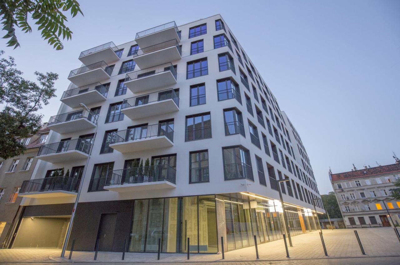 Nowoczesny budynek apartamentowy z balkonami, widok z dołu, szklane witryny na parterze, oświetlenie uliczne, błękitne niebo w tle.