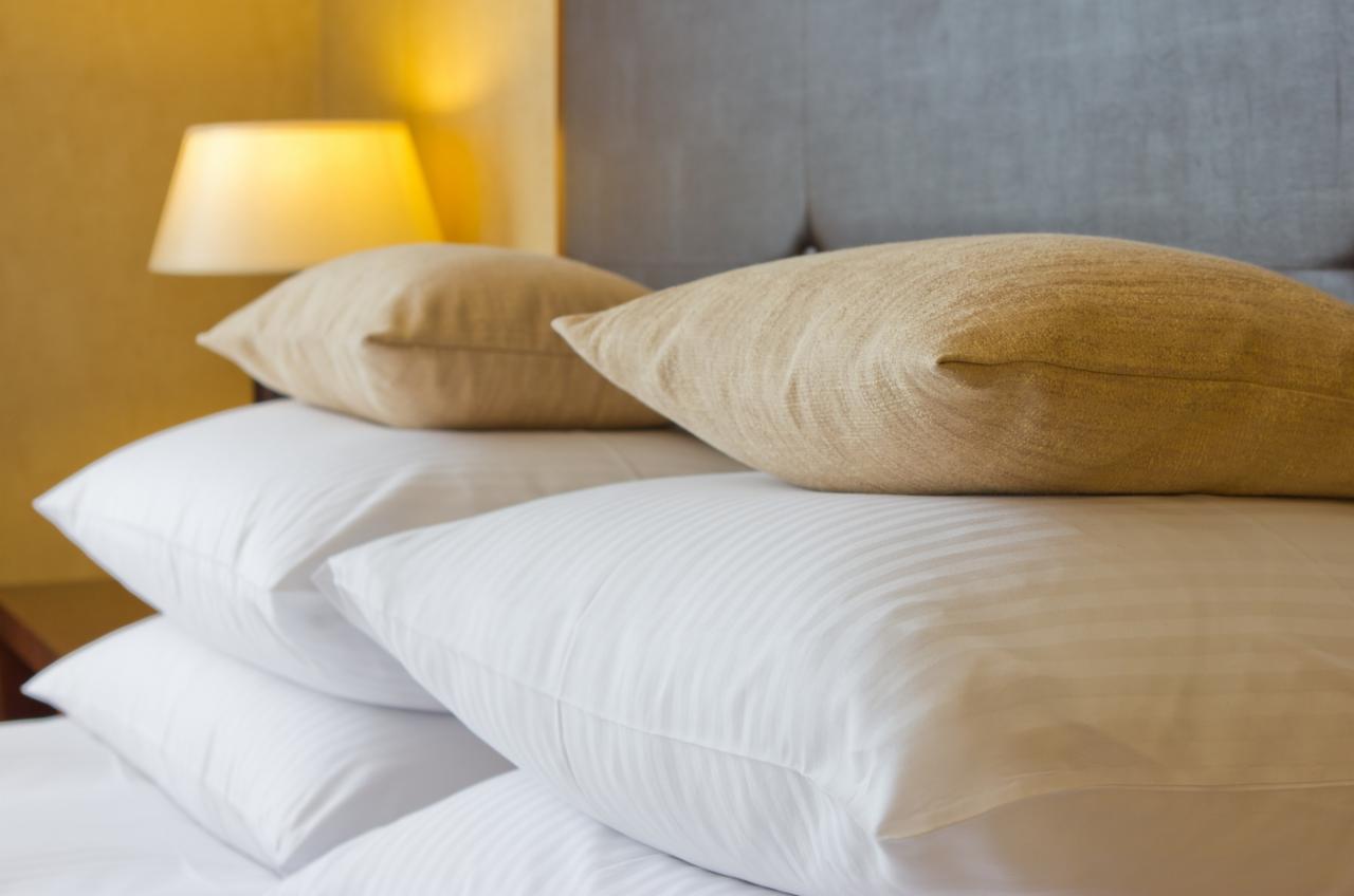 Stos białych poduszek z delikatnym paskowaniem i dwie beżowe poduszki na wierzchu, oświetlone ciepłym światłem lampki nocnej.