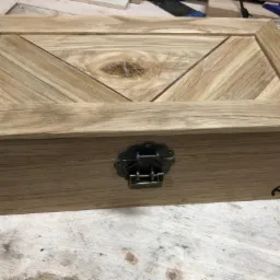 Drewniana skrzynka z dekoracyjnym wiekiem ułożonym z drewnianych listewek w jodełkę, zamknięta na metalowy zamek ze zdobieniami, leżąca na warsztatowym stole.