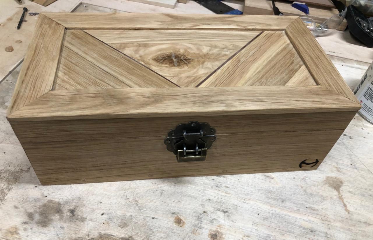 Drewniana skrzynka z dekoracyjnym wiekiem ułożonym z drewnianych listewek w jodełkę, zamknięta na metalowy zamek ze zdobieniami, leżąca na warsztatowym stole.