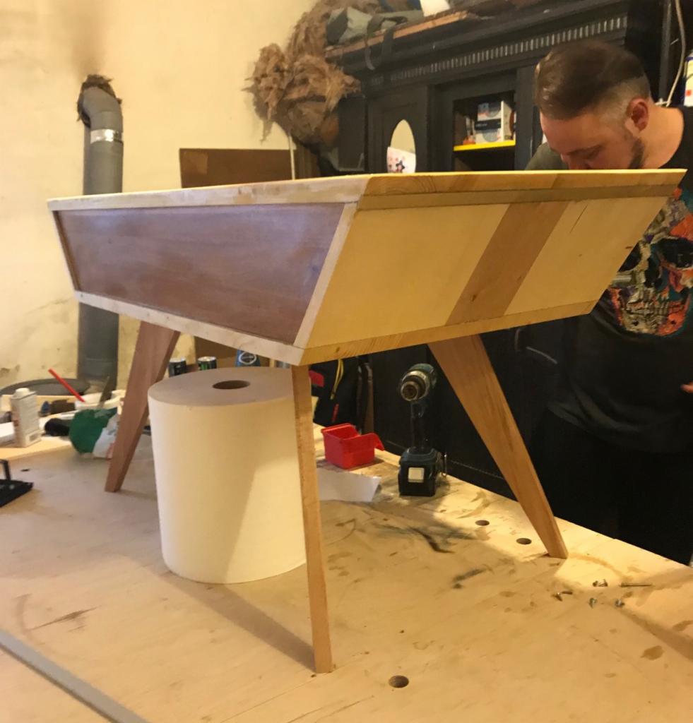 Drewniana skrzynia na nogach w trakcie montażu, widoczny warsztat, narzędzia i majsterkowicz w tle.