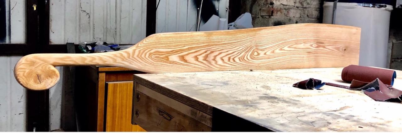 Element drewniany z zakręconym uchwytem, prawdopodobnie fragment balustrady lub poręczy, w trakcie obróbki szlifierskiej na warsztatowym stole, widoczne ślady pyłu i narzędzia do szlifowania.