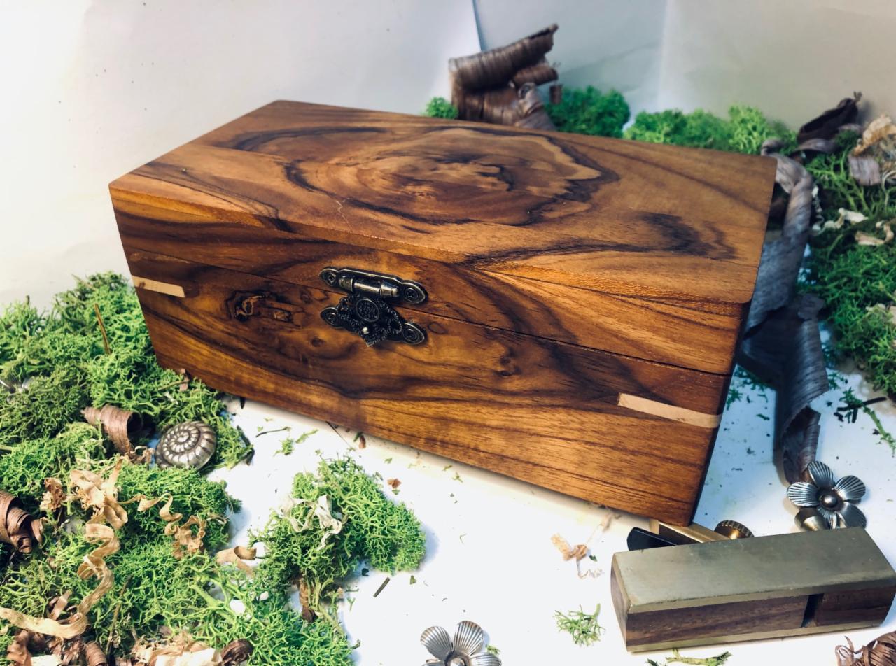 Drewniana szkatułka z ozdobnym zapięciem, wykonana z egzotycznego drewna o wyraźnym usłojeniu, prezentowana na tle mchu i wiórów, obok strug do drewna.