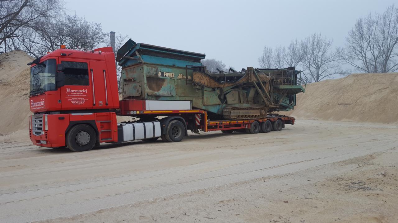 Czerwona ciężarówka z naczepą transportuje dużą, używaną przesiewarkę do kruszyw Power Screen, widoczne ślady opon na piaszczystym podłożu, hałdy piasku w tle.