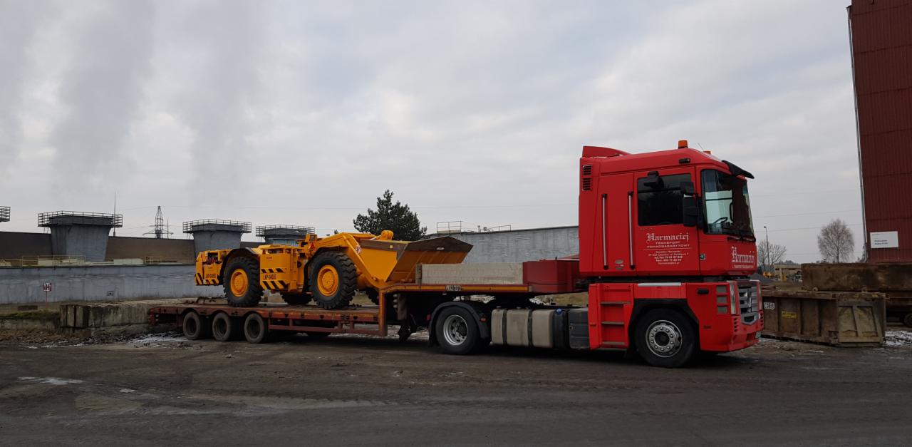 Czerwona ciężarówka transportuje żółtą maszynę górniczą na niskopodwoziowej platformie, widoczne elementy konstrukcyjne w tle.