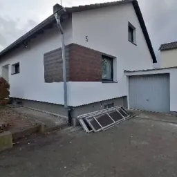 Biały dom jednorodzinny z garażem, częściowo ocieplony brązowymi panelami, z numerem '5' na ścianie, okna w czarnych ramach, widoczne okna piwniczne i elementy rusztowania.