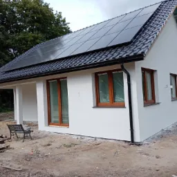 Nowoczesny, biały dom jednorodzinny z brązowymi oknami i panelami słonecznymi na czarnym dachu, widok z boku, teren wokół domu w trakcie prac wykończeniowych.