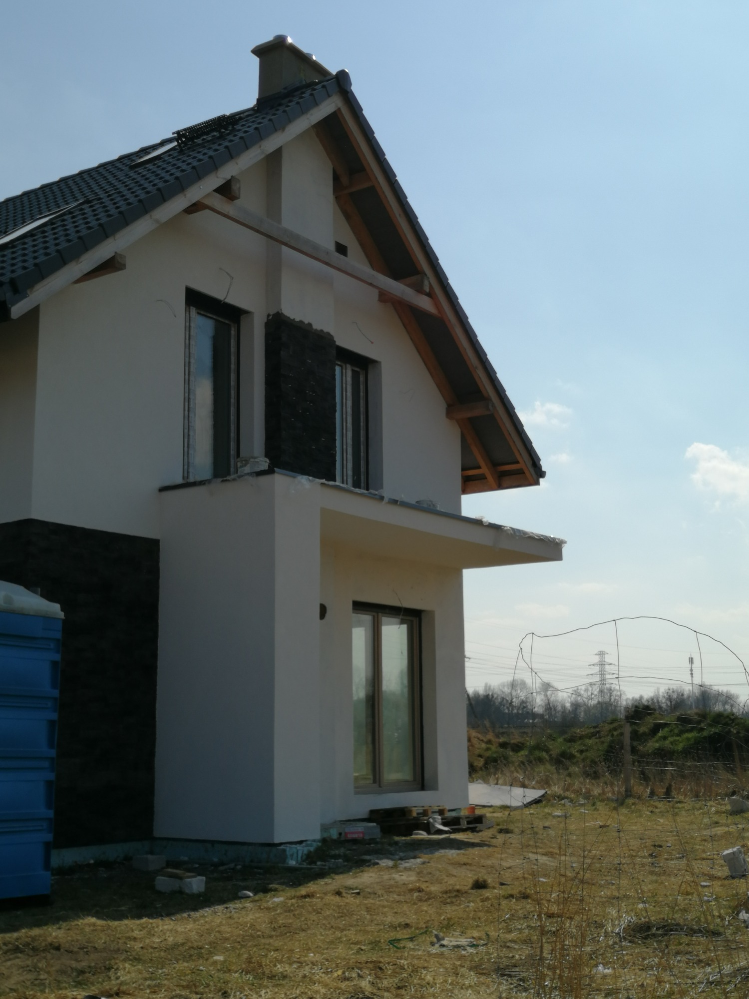 Niedokończony dom jednorodzinny z fragmentami elewacji w trakcie wykończenia, widoczne drewniane elementy konstrukcyjne dachu i niebieska toaleta przenośna.