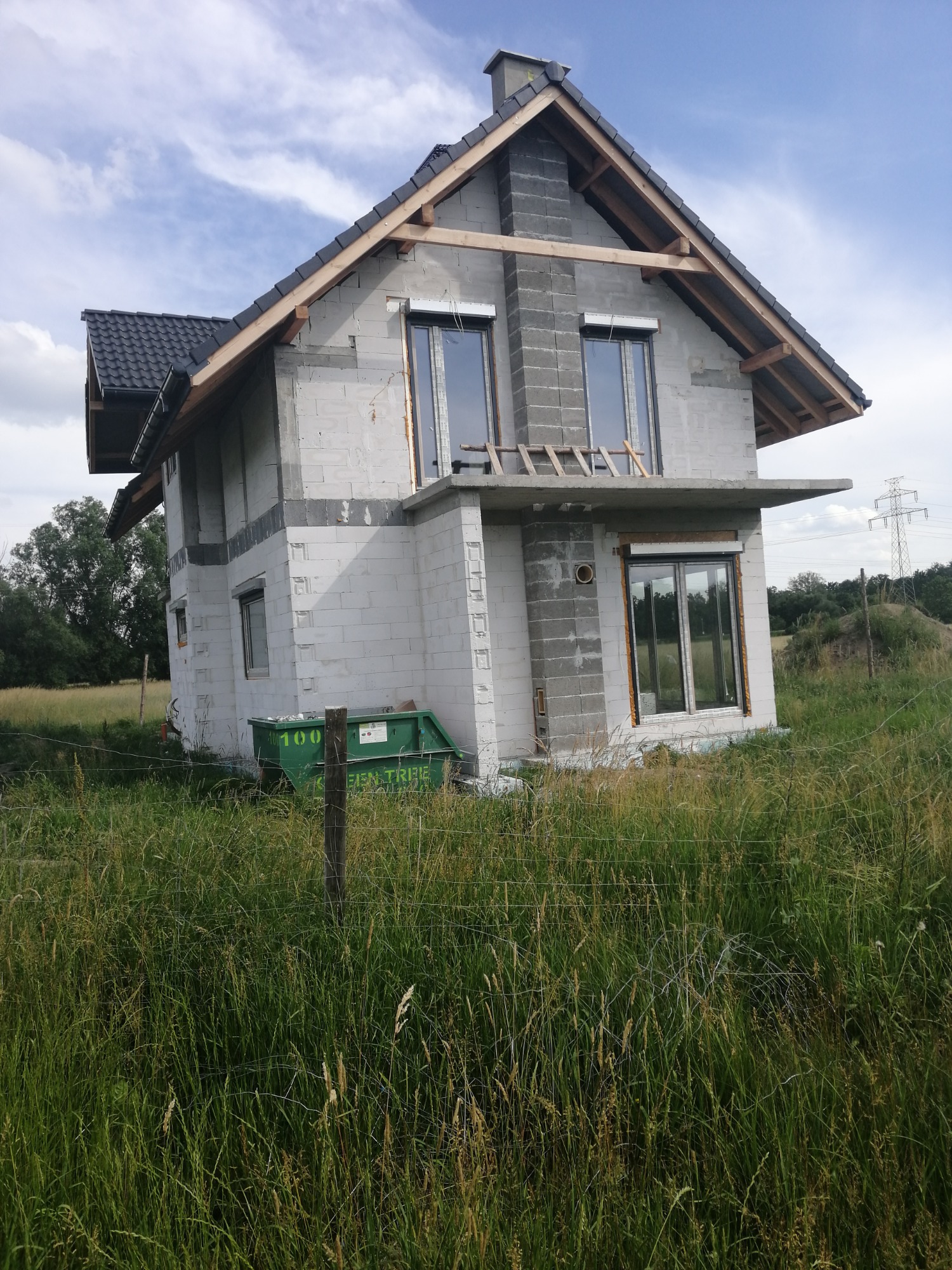 Niedokończony dom z szarego bloczka, z widoczną konstrukcją dachu i oknami z roletami, na tle łąki z wysoką trawą i niebieskiego nieba.