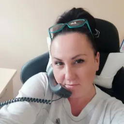 Kobieta z okularami na głowie siedzi na krześle biurowym, trzymając słuchawkę telefonu przy uchu i spoglądając w obiektyw.