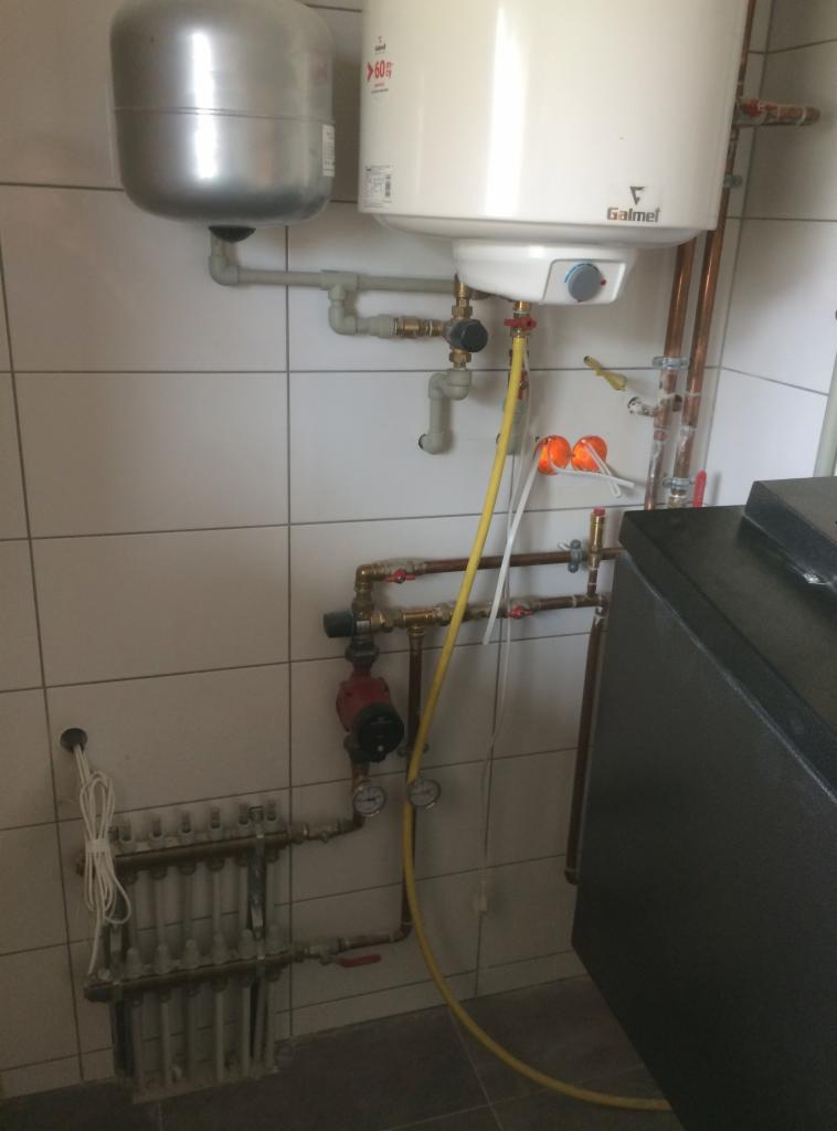 Bojler Galmet z podłączonymi rurami miedzianymi i plastikowymi, widoczny również zawór, pompa obiegowa i kolektor rozdzielacza, całość zamontowana na ścianie z białych płytek.