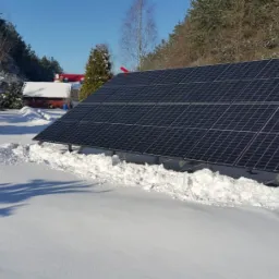 Panel słoneczny zainstalowany na śniegu w zimowy dzień, widoczny dom i drzewa w tle.
